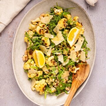 Roasted cauliflower salad on a platter with wooden spoon next to linen and garlic bulb.