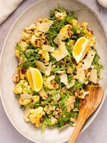 Roasted cauliflower salad on a platter with wooden spoon next to linen and garlic bulb.