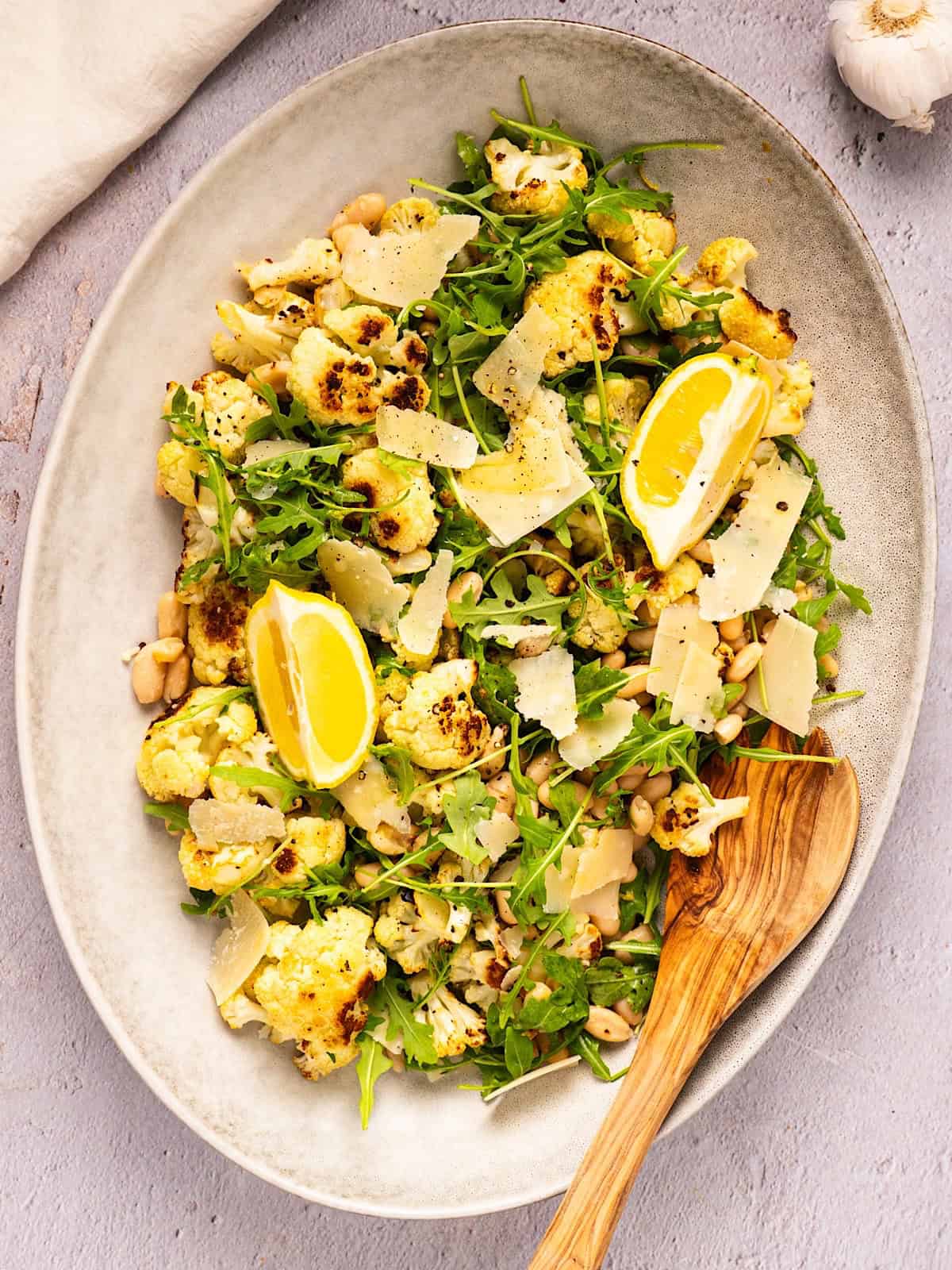 Roasted cauliflower salad on a platter with wooden spoon next to linen and garlic bulb.
