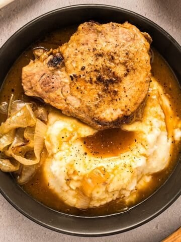 A pork chop layered over mashed potatoes with onion gravy and a side of onions in a black bowl.