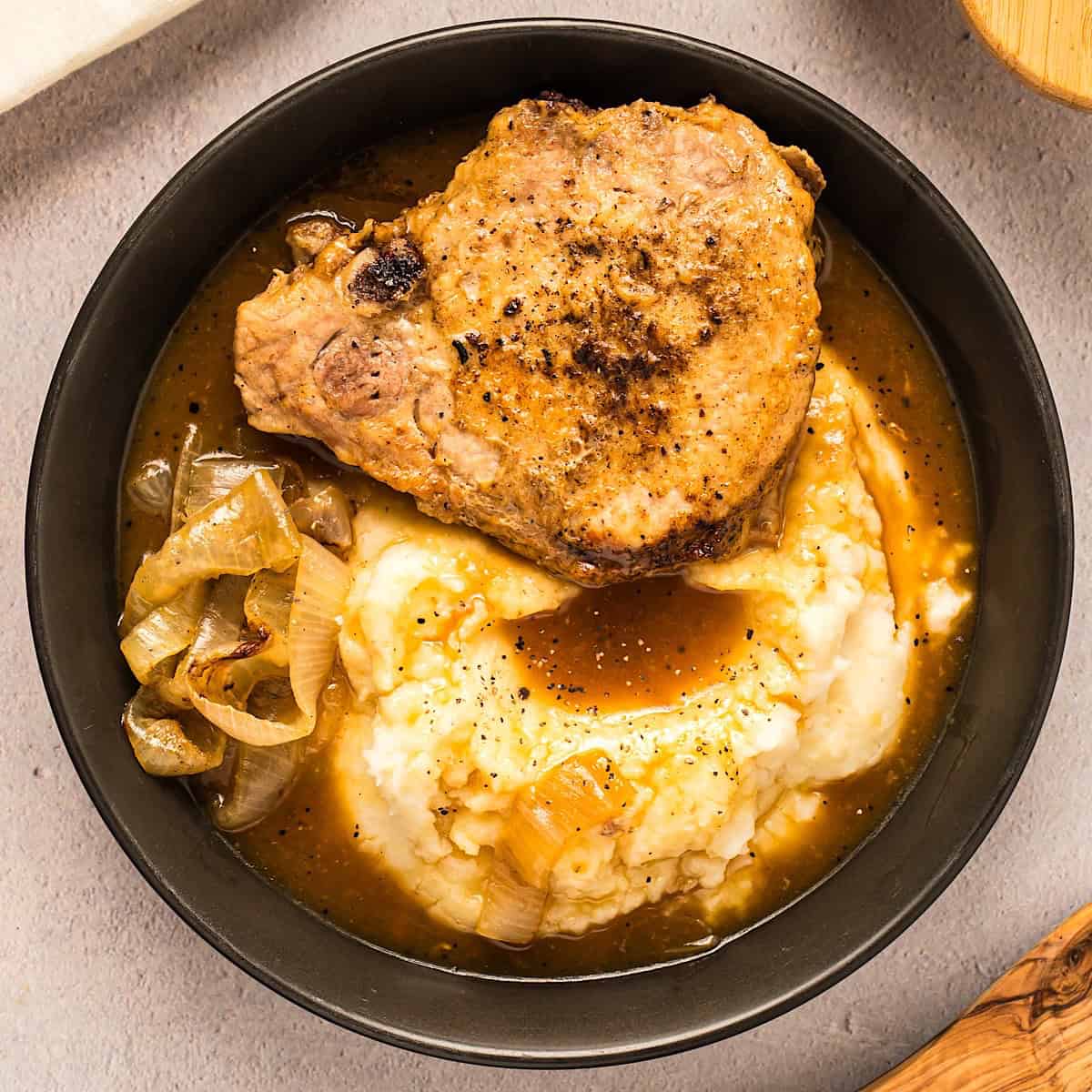 A pork chop layered over mashed potatoes with onion gravy and a side of onions in a black bowl.