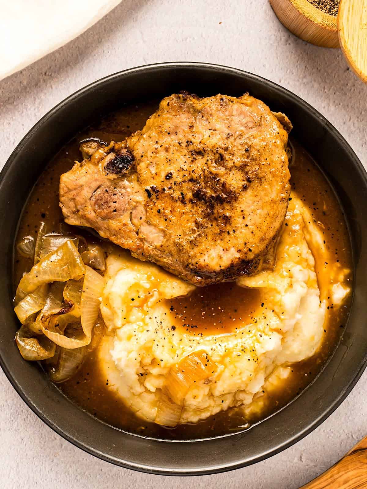 A pork chop layered over mashed potatoes with onion gravy and a side of onions in a black bowl.
