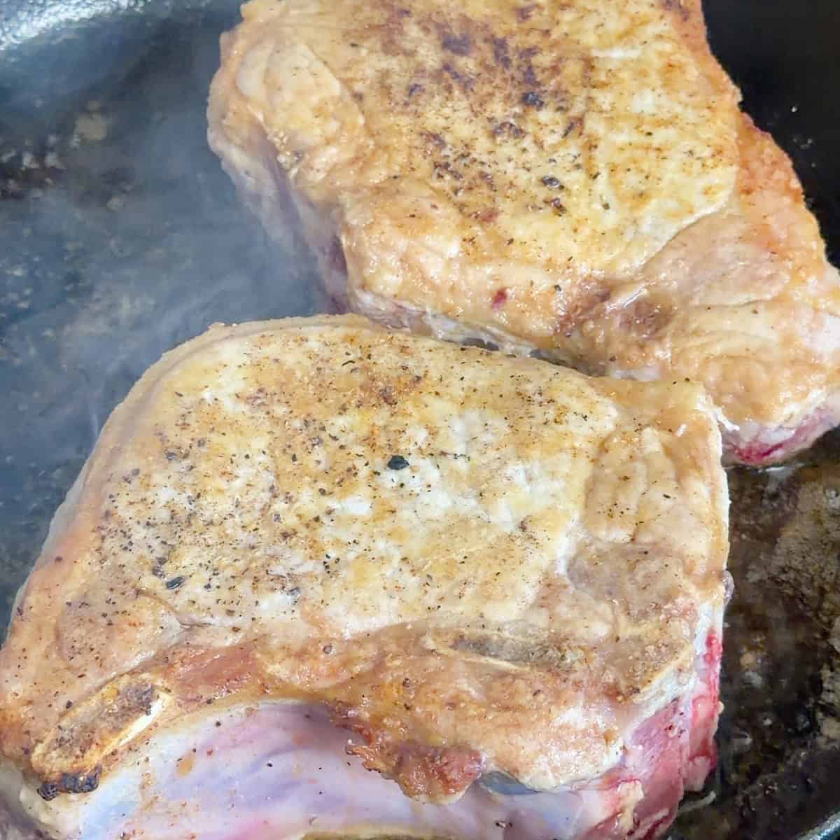 Two pork chops searing to golden brown in a cast iron skillet.