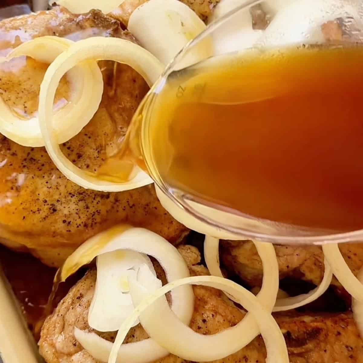 Pouring the broth around the seared pork chops and onions in a casserole dish.
