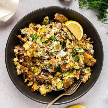 Crispy smashed potato salad in a large serving bowl with fork and lemon wedge, garlic bulb and linen in background.
