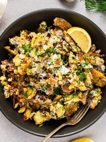 Crispy smashed potato salad in a large serving bowl with fork and lemon wedge, garlic bulb and linen in background.