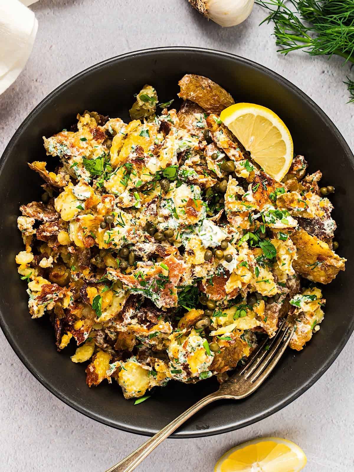 Crispy smashed potato salad in a large serving bowl with fork and lemon wedge, garlic bulb and linen in background.