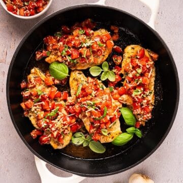 Bruschetta chicken in a skillet with fresh basil leaves near a garlic bulb and small bowl of topping.