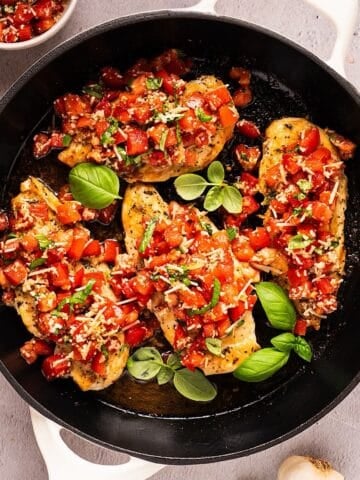 Bruschetta chicken in a skillet with fresh basil leaves near a garlic bulb and small bowl of topping.