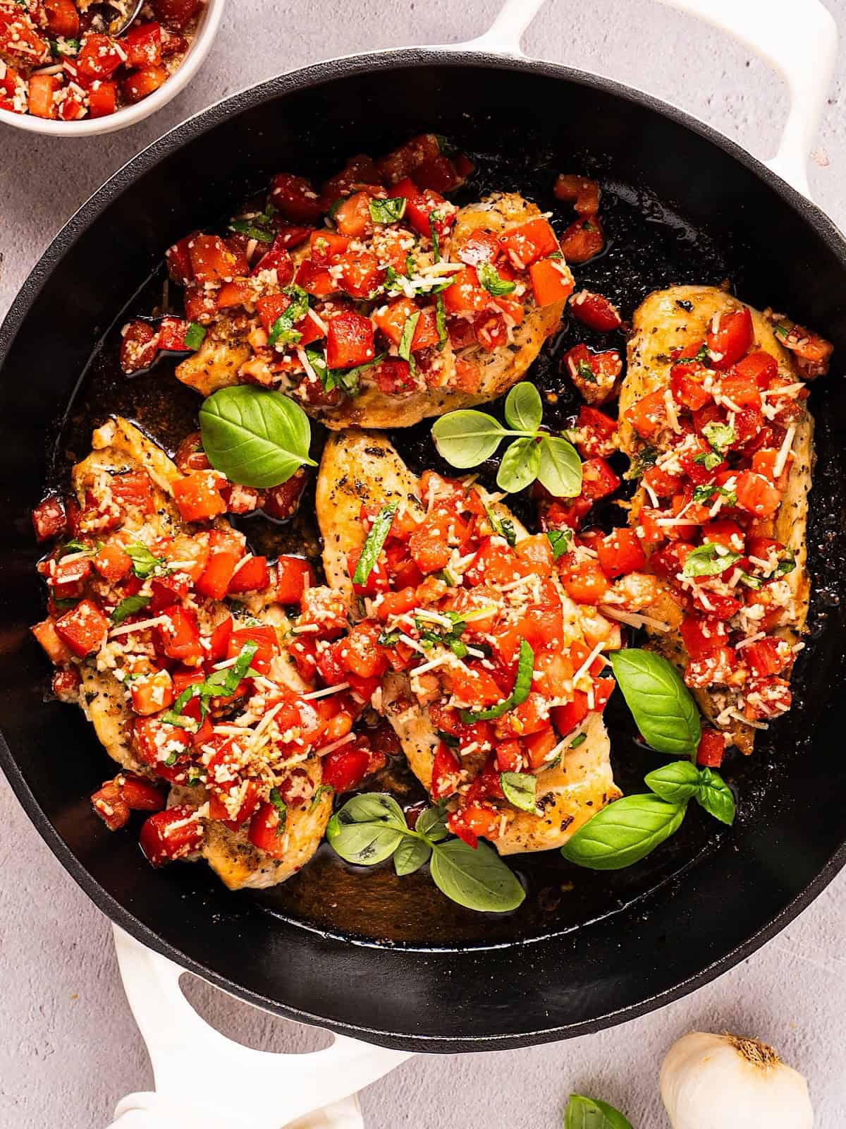 Bruschetta chicken in a skillet with fresh basil leaves near a garlic bulb and more basil.