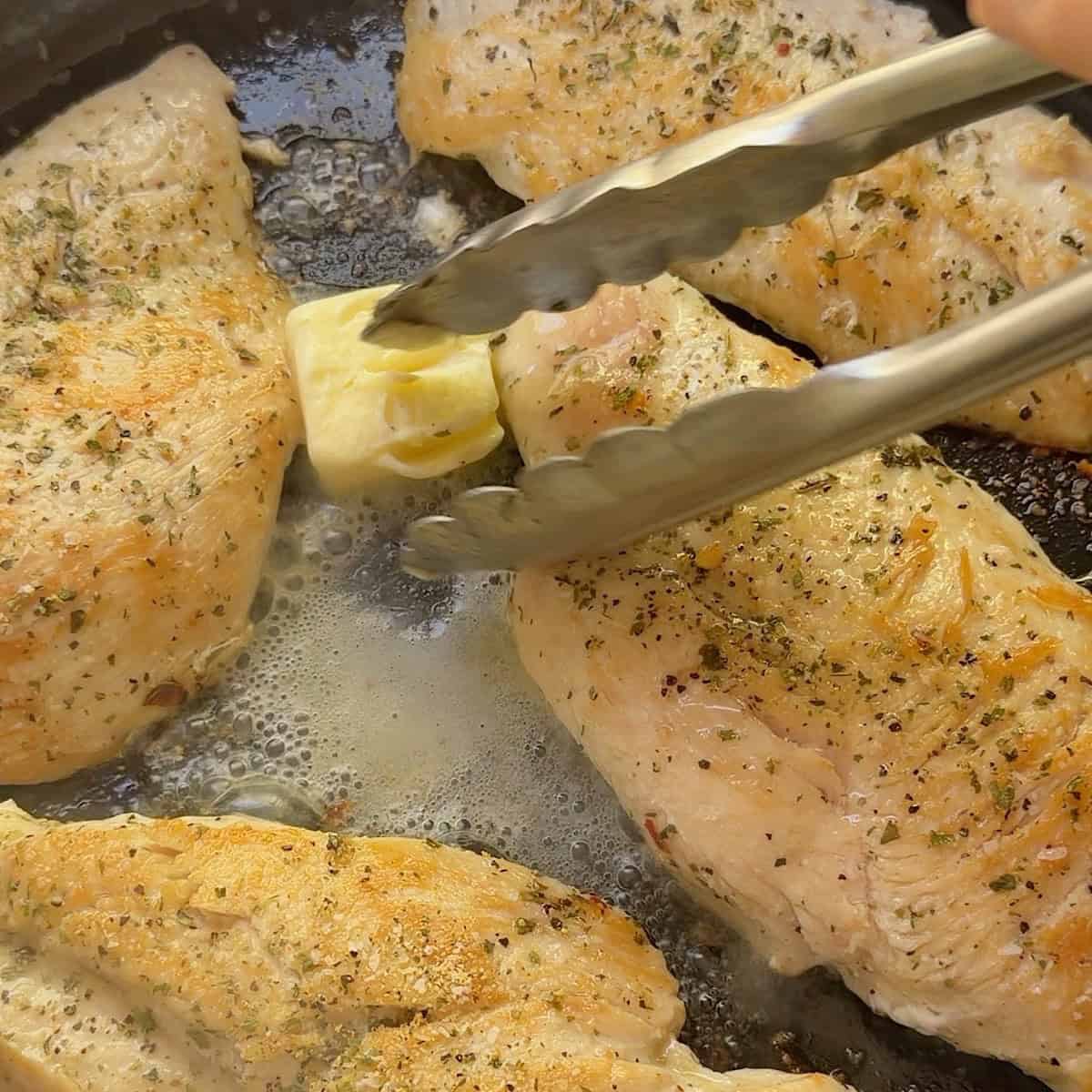 Adding butter to the skillet with tongs ready to flip chicken.