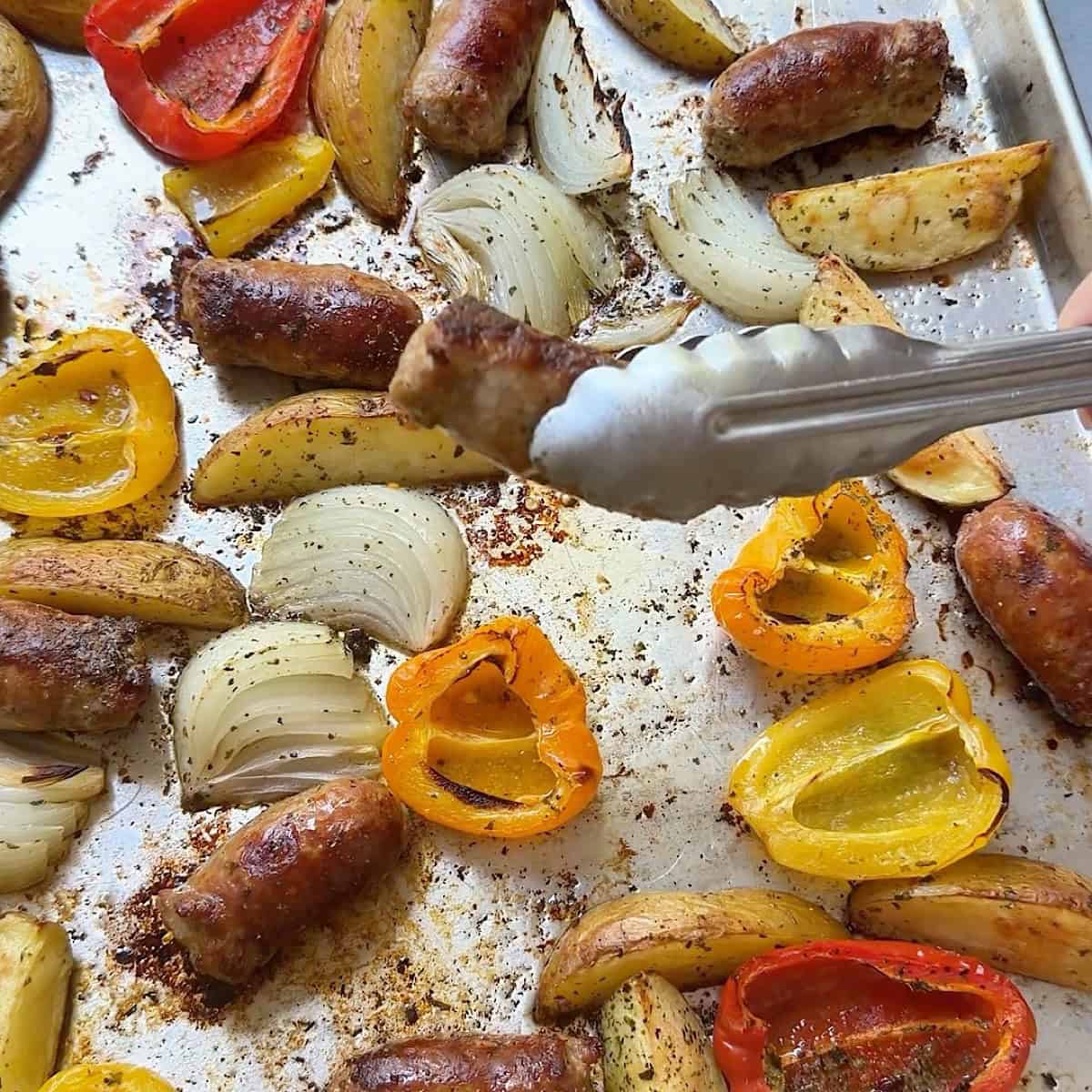 Fully baked sheet pan sausage and potatoes with crispy brown edges and tongs holding sausage.
