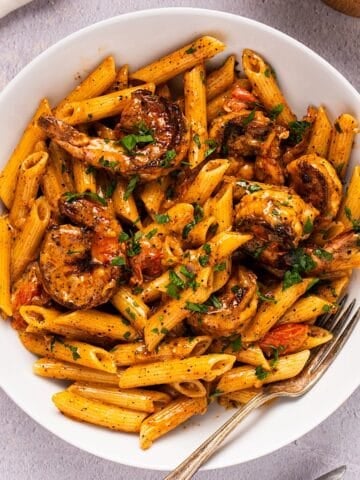 Creamy blackened shrimp pasta with garnish in a white bowl plated next to linen and wooden box.