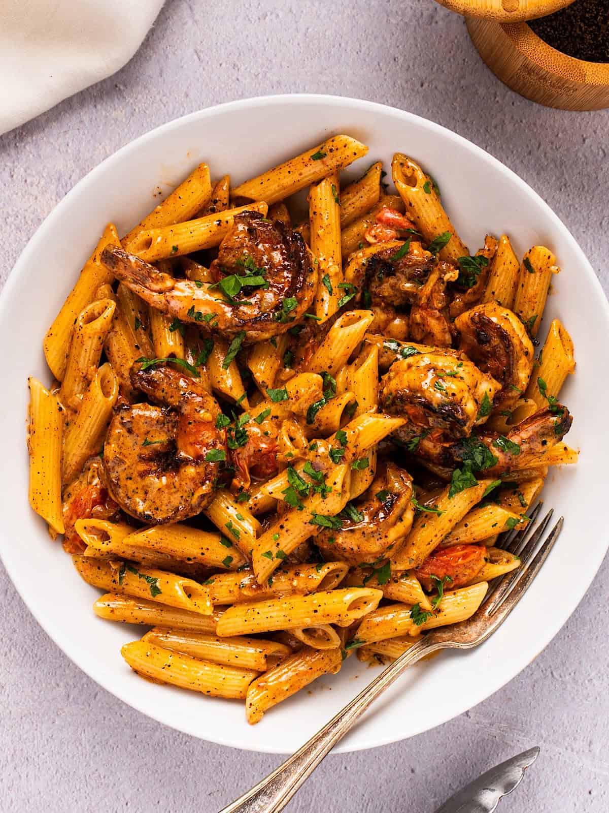 Creamy blackened shrimp pasta with garnish in a white bowl plated next to linen and wooden box.