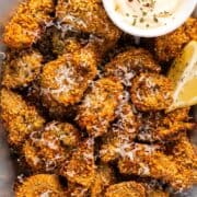 A platter of crispy artichoke hearts with lemon wedges, parmesan garnish, and a small bowl of garlic aioli.