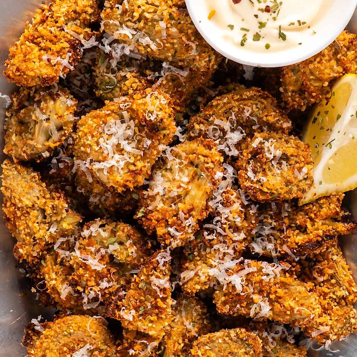 A platter of crispy artichoke hearts with lemon wedges, parmesan garnish, and a small bowl of garlic aioli.
