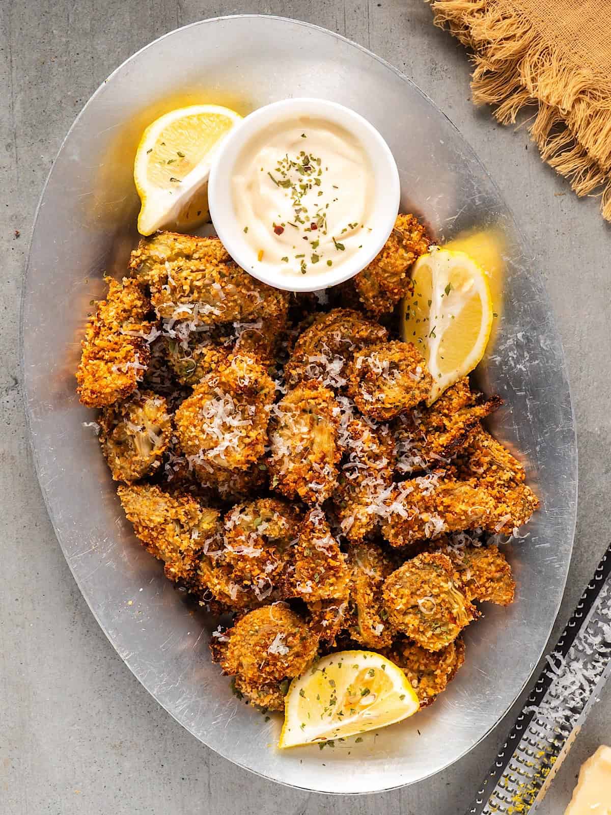 A platter of crispy artichoke hearts with lemon wedges, parmesan garnish, and a small bowl of garlic aioli.