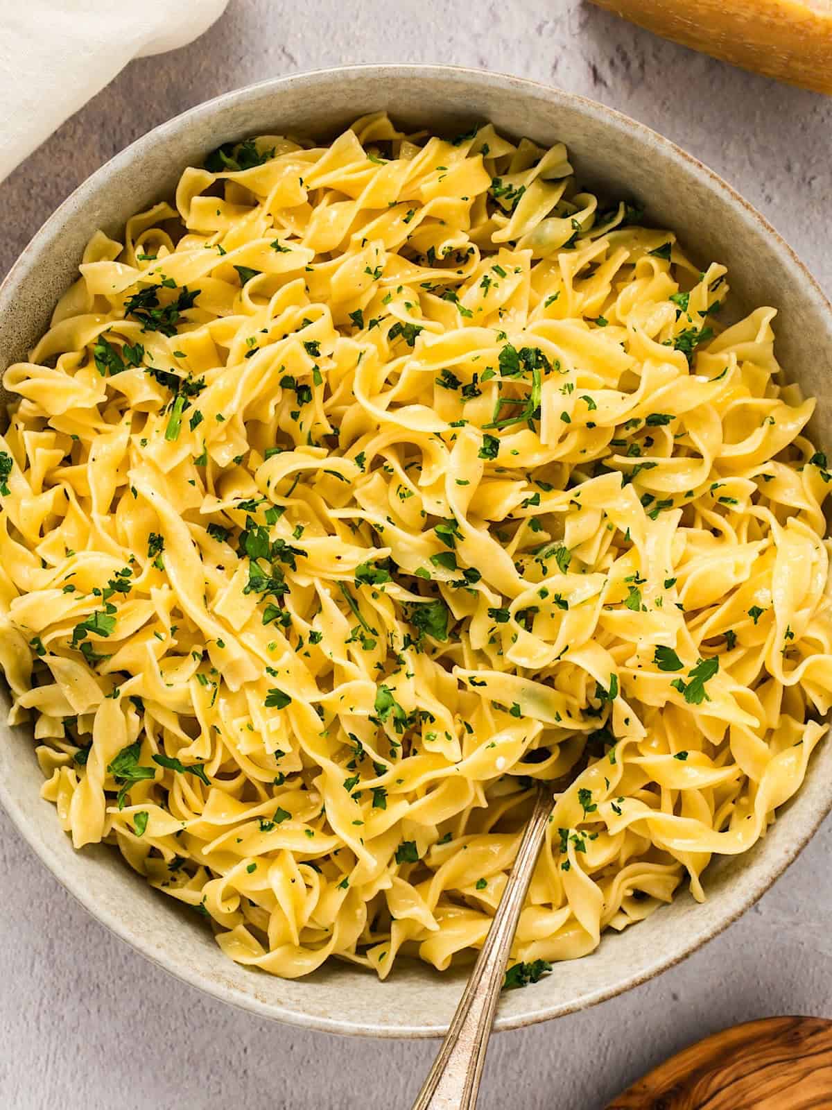 A wide bowl of buttered egg noodles topped with parsley plated next to parmesan, a linen, and wooden spoon.