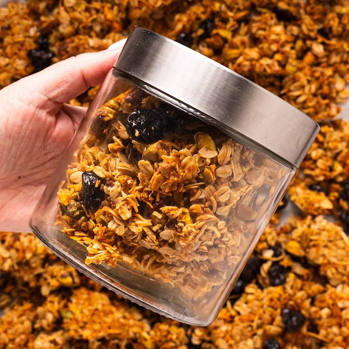 Holding a jar of homemade honey granola over the sheet pan of granola.