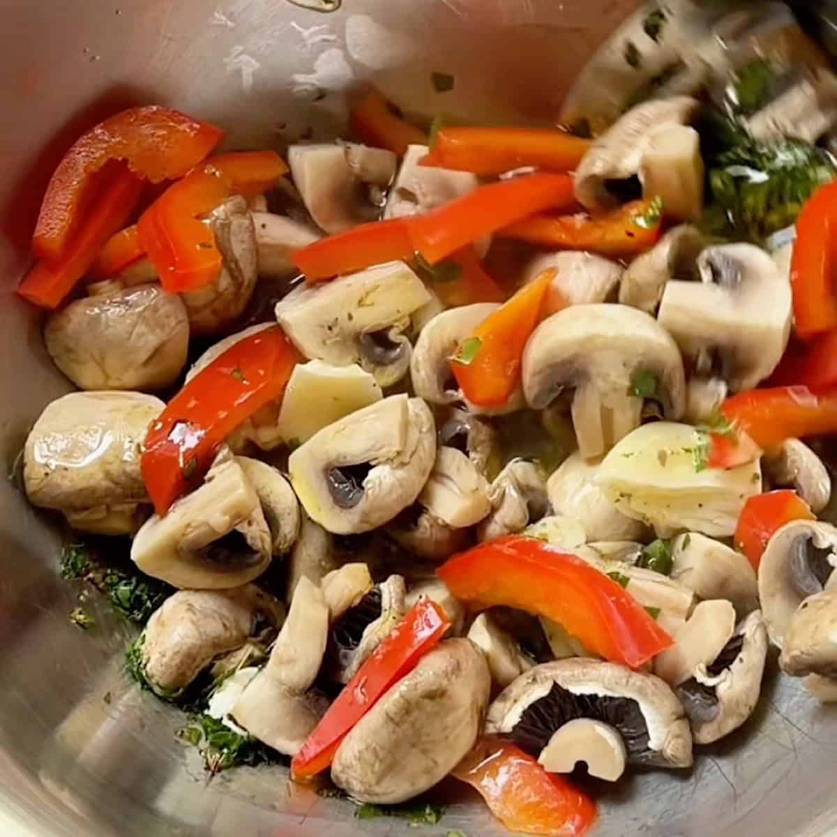Mushrooms and peppers being tossed in seasoned olive in a mixing bowl.