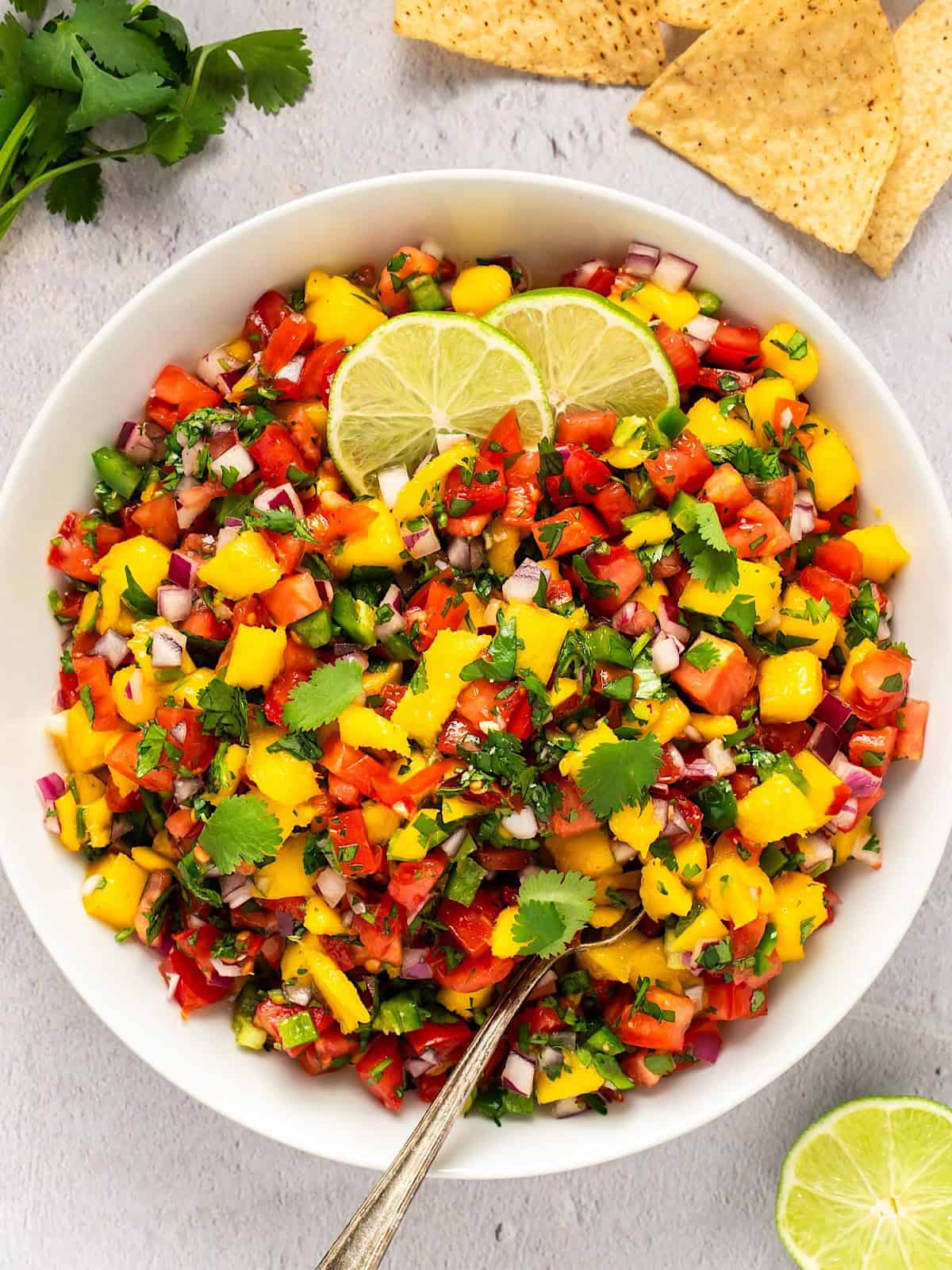 Mango pico de gallo in a white bowl next to lime, tortilla chips, and cilantro.