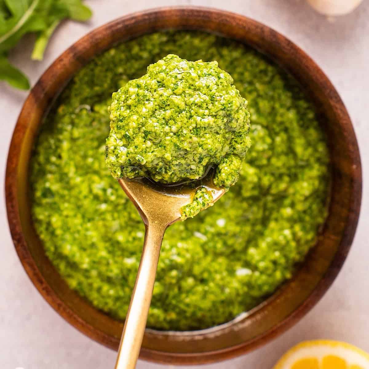 A gold spoonful of basil arugula pesto over a wooden bowl of pesto.