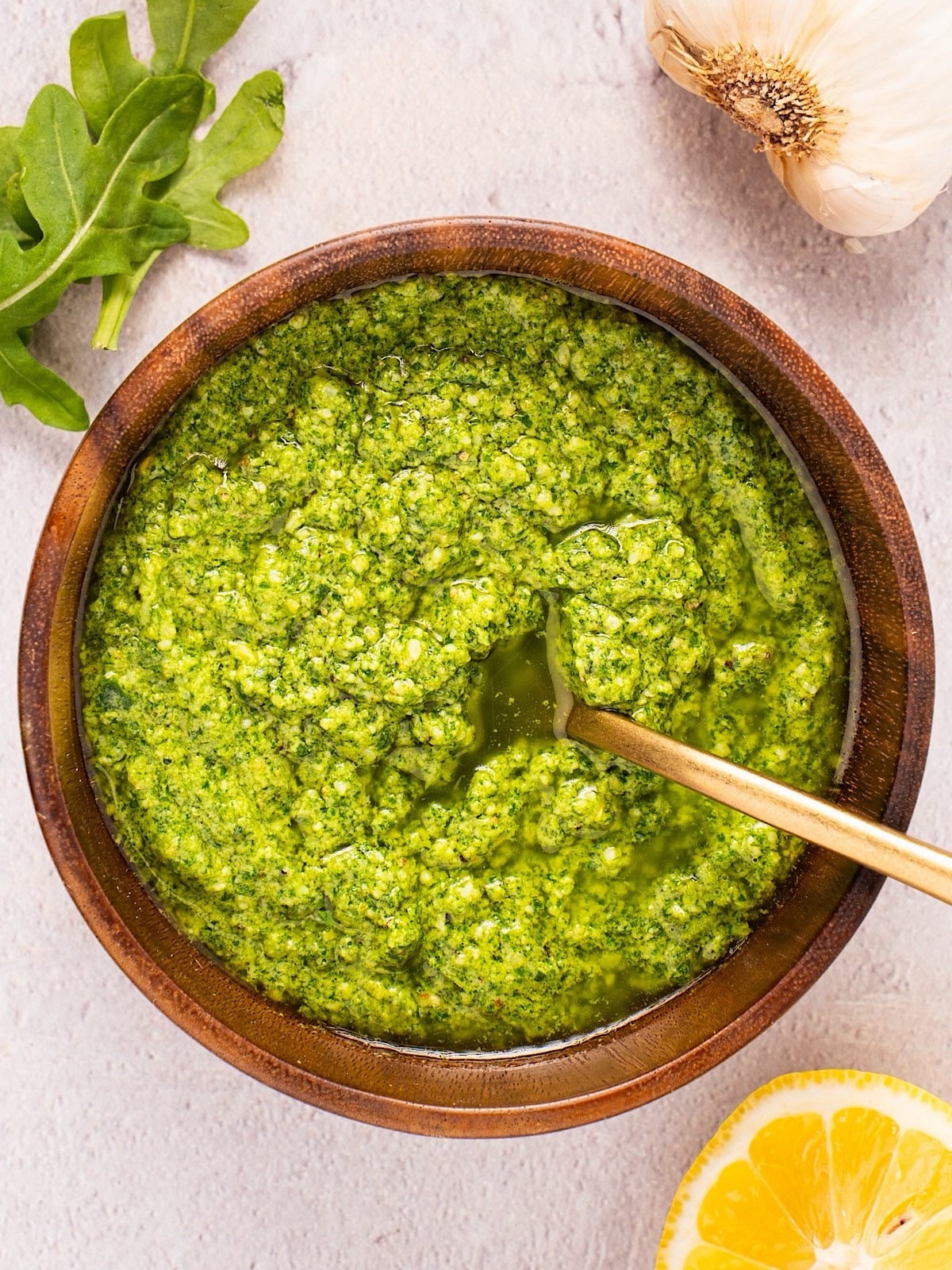 A wooden bowl of basil arugula pesto with gold spoon next to a garlic bulb and arugula leaves.