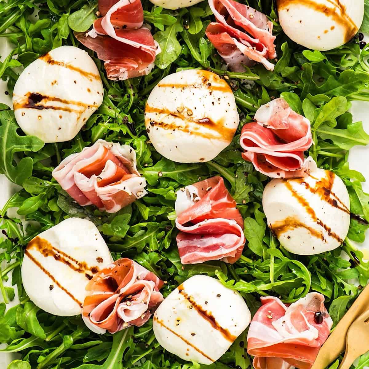 Burrata and prosciutto on a platter of arugula dressed with olive oil and balsamic glaze.