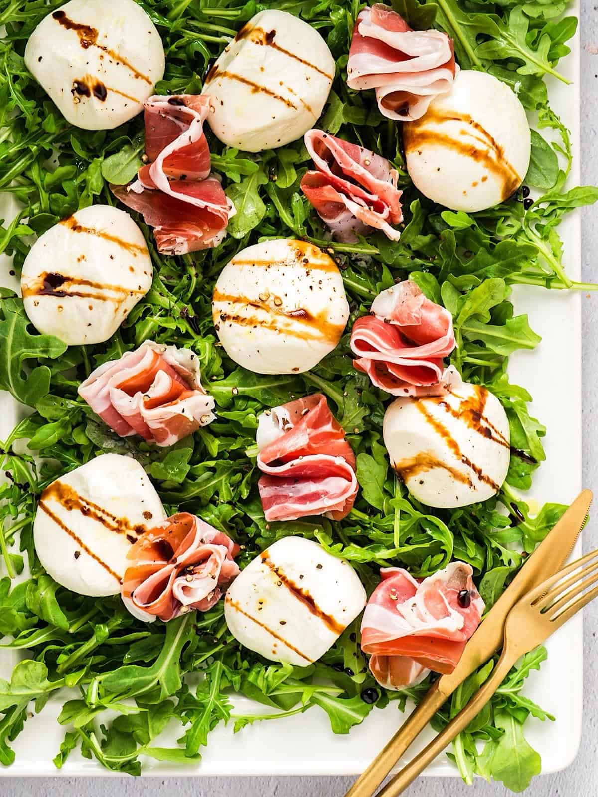 Balsamic glazed burrata and prosciutto on a platter of arugula with gold fork and knife.