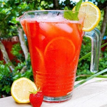 Fresh homemade strawberry lemonade in a pitcher on a deck with lemon and mint garnish.