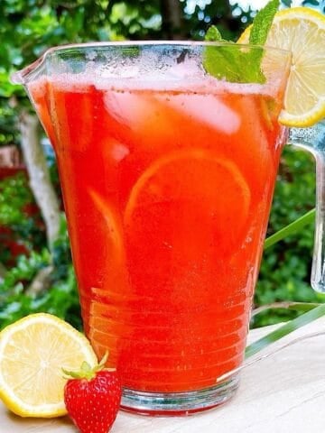Fresh homemade strawberry lemonade in a pitcher on a deck with lemon and mint garnish.