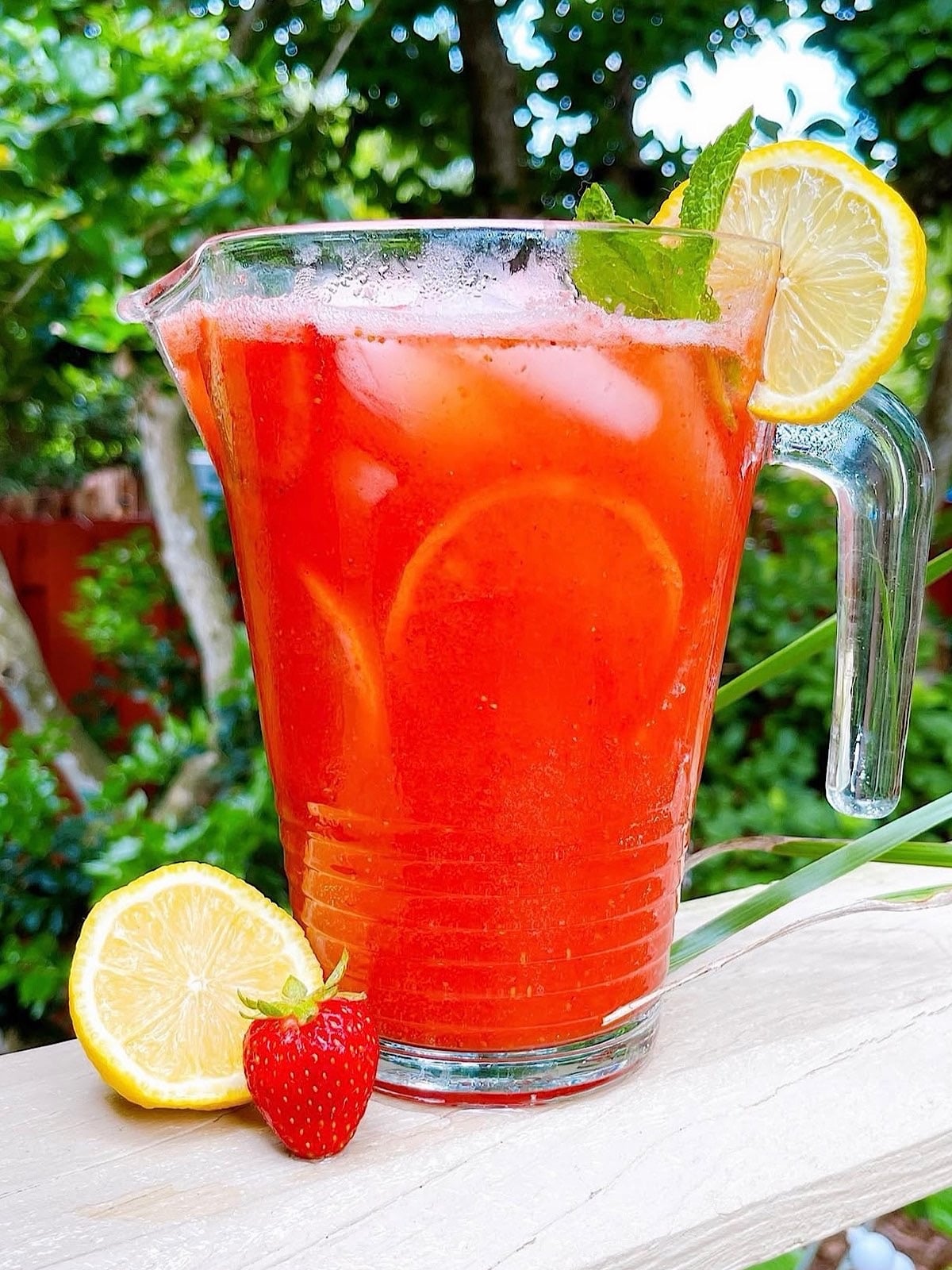 Fresh homemade strawberry lemonade in a pitcher on a deck with lemon and mint garnish.