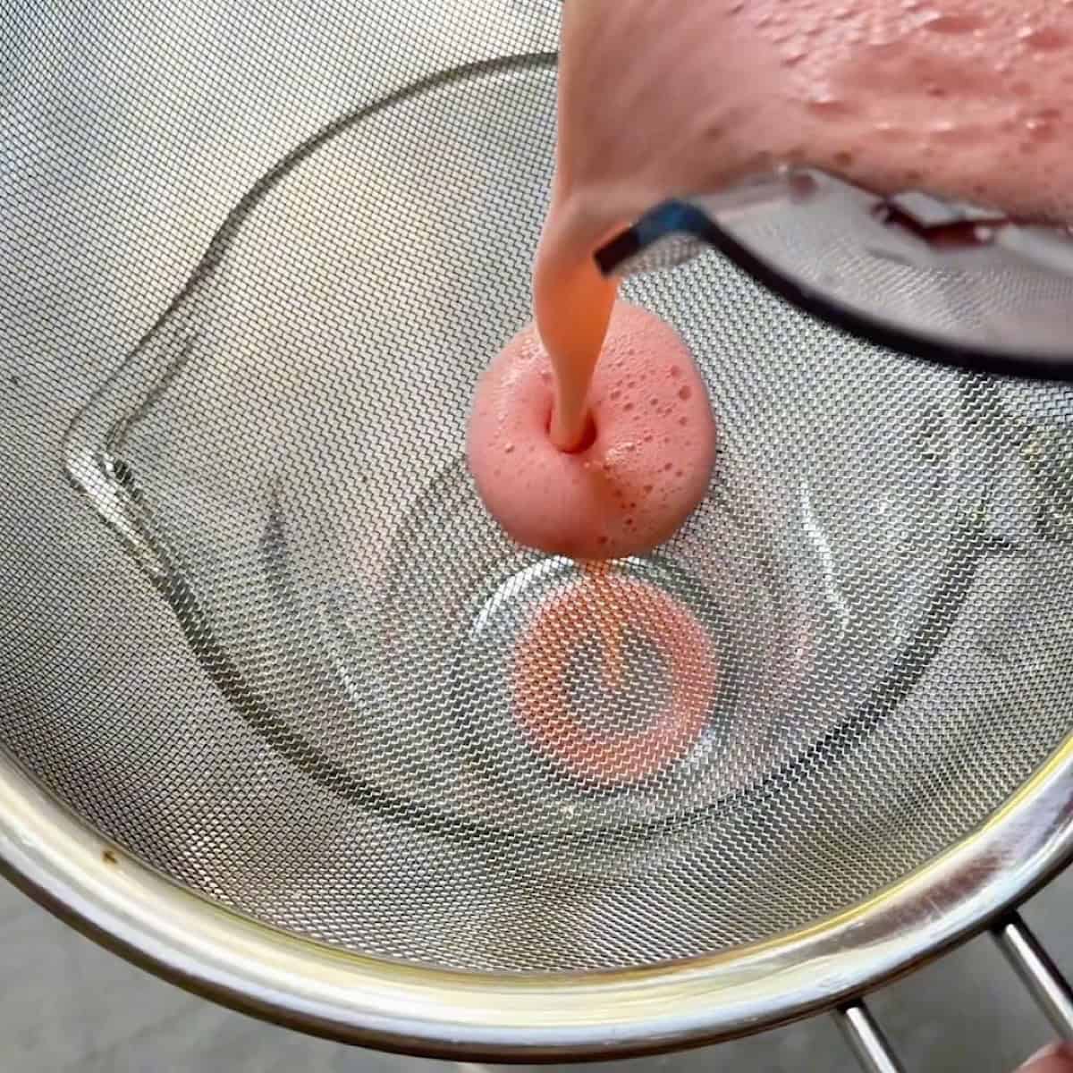 Strawberry lemonade being poured through a strainer into a pitcher.
