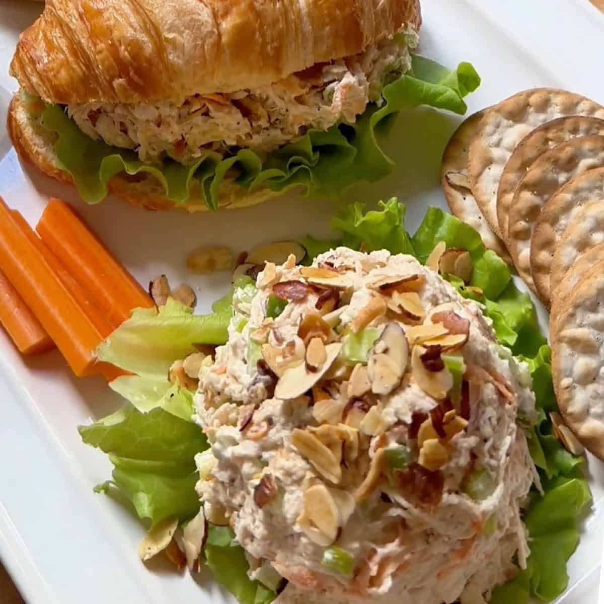 A platter with a mound of honey chicken salad, two croissant sandwiches, carrot sticks, crackers, and a side of honey for dipping.