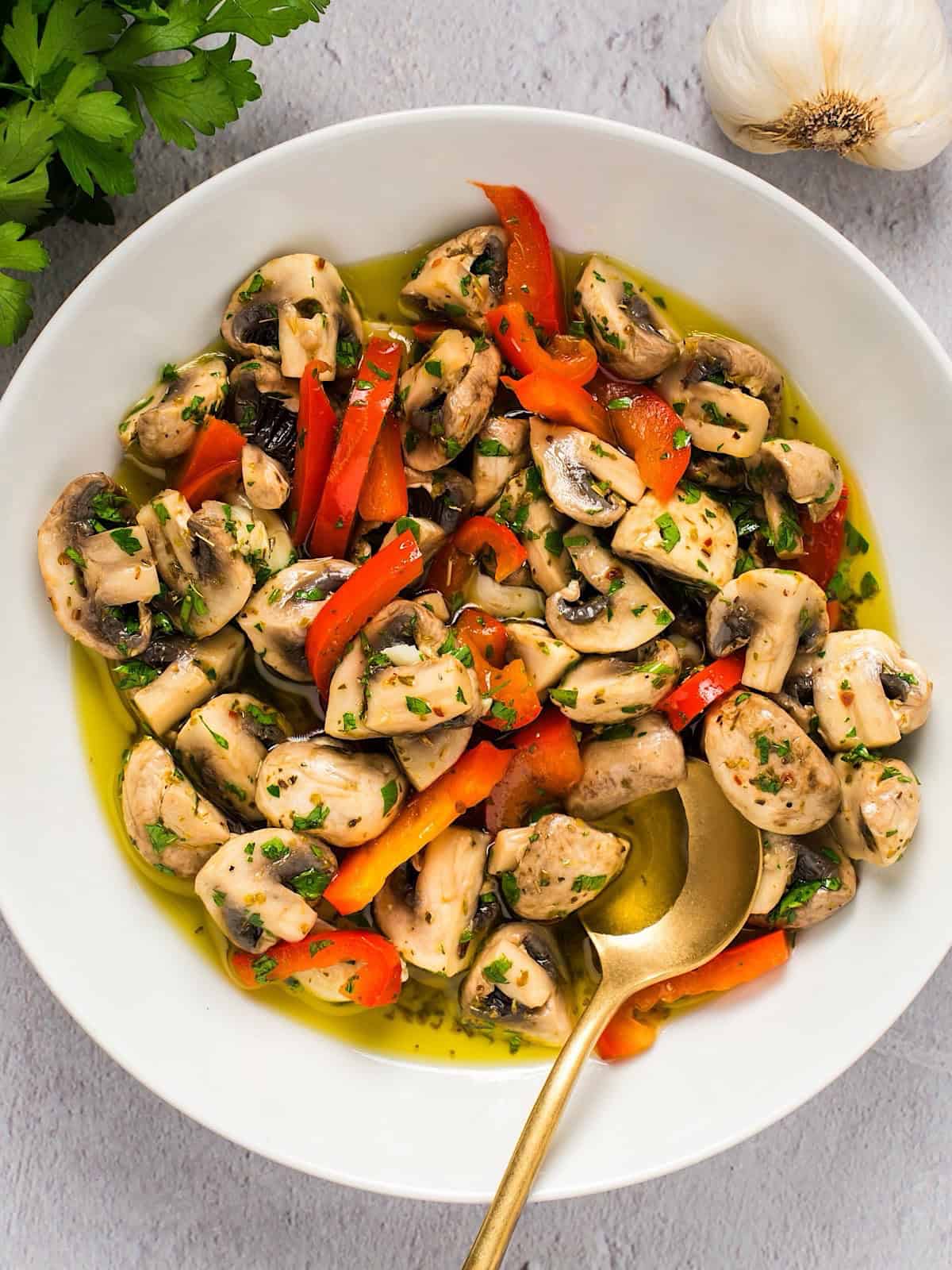 Italian marinated mushrooms in a white bowl with garlic, parsley, and gold spoon for composition.