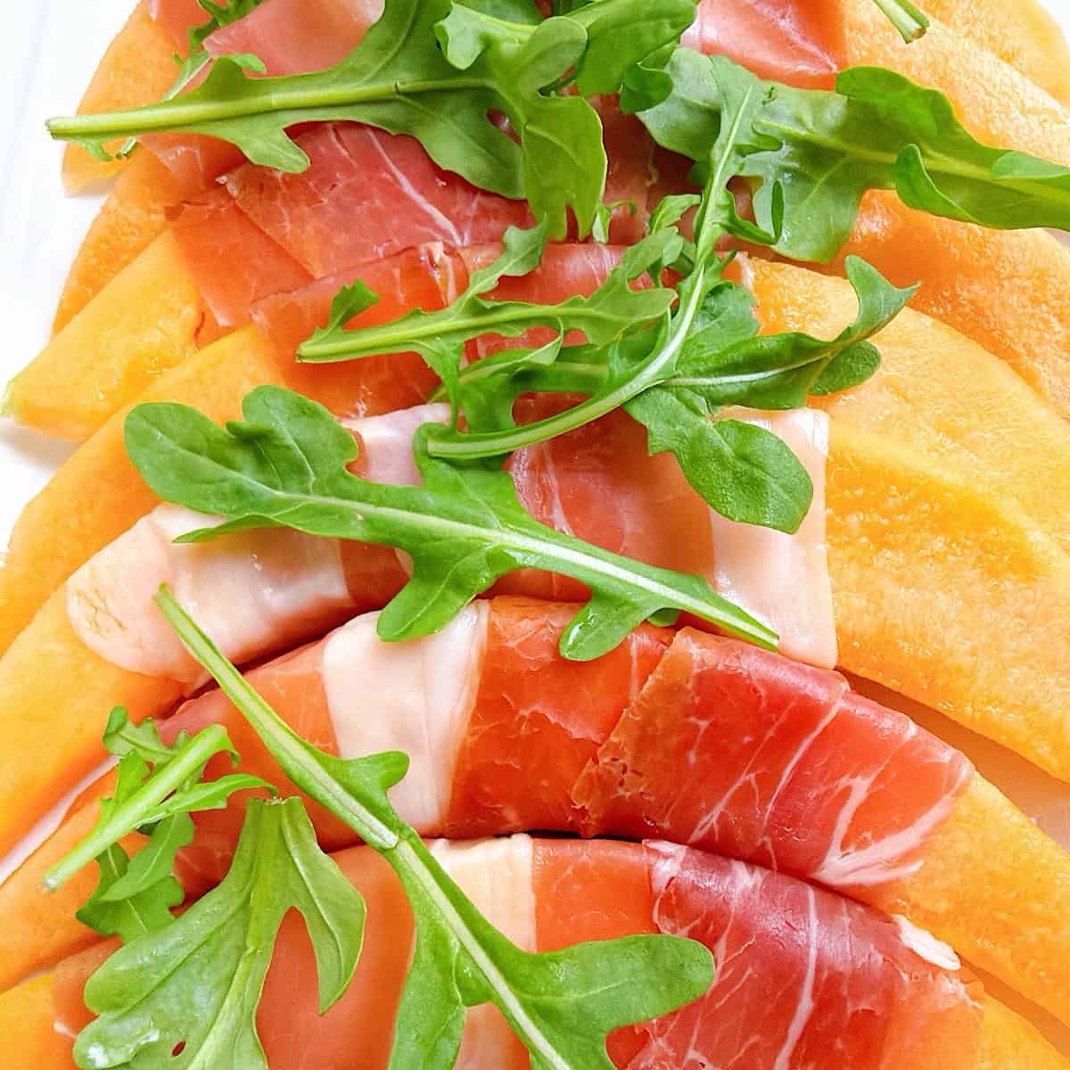 Prosciutto wrapped cantaloupe arranged on a platter with arugula scattered on top.