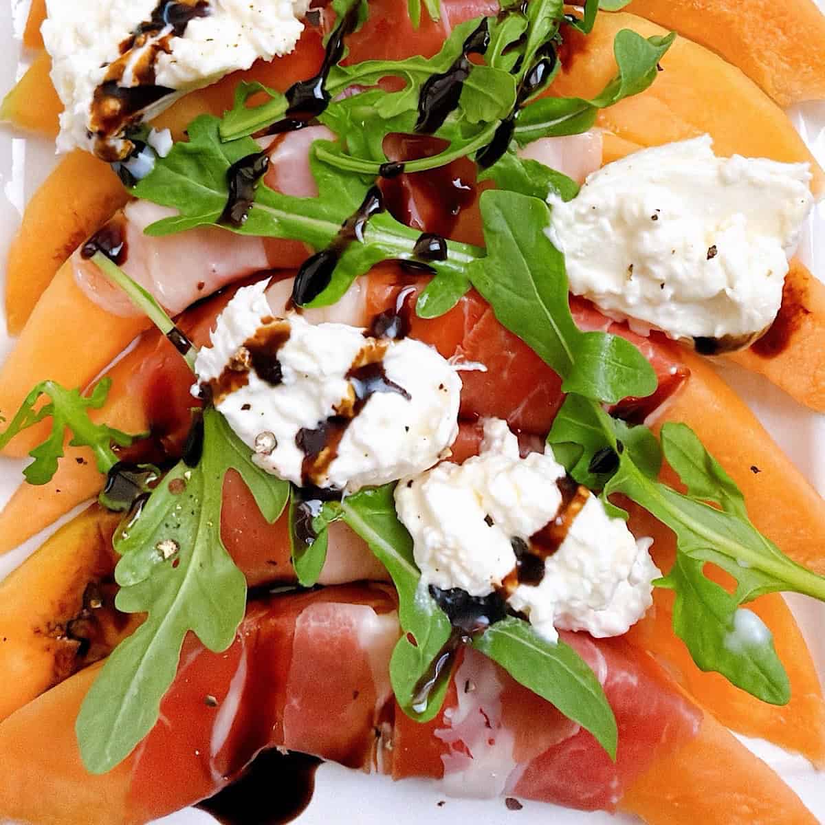 Prosciutto wrapped cantaloupe on a platter with arugula, burrata cheese, and balsamic glaze.