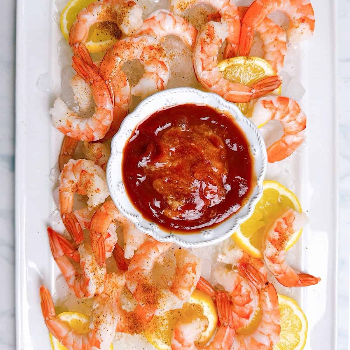 Shrimp cocktail platter on a tray with crushed ice, lemon slices, and a bowl of cocktail sauce in the center.