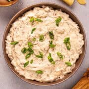 Caramelized onion dip with green onion garnish in a bowl plated next to chips and wooden spoon.