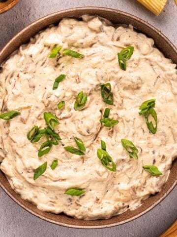 Caramelized onion dip with green onion garnish in a bowl plated next to chips and wooden spoon.