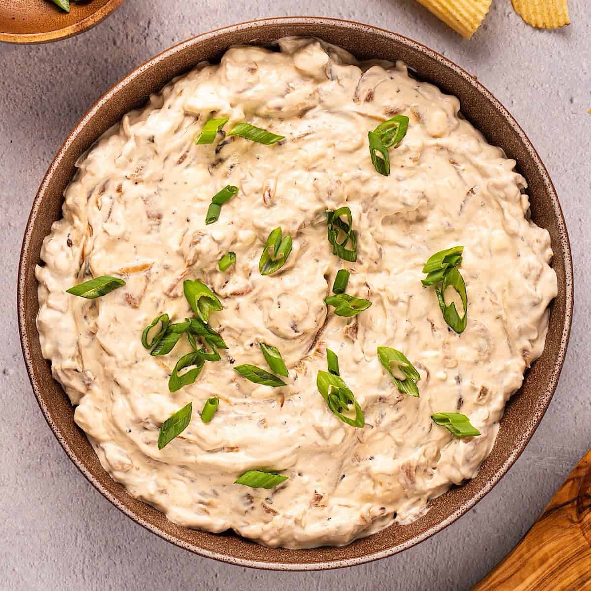 Caramelized onion dip with green onion garnish in a bowl plated next to chips and wooden spoon.