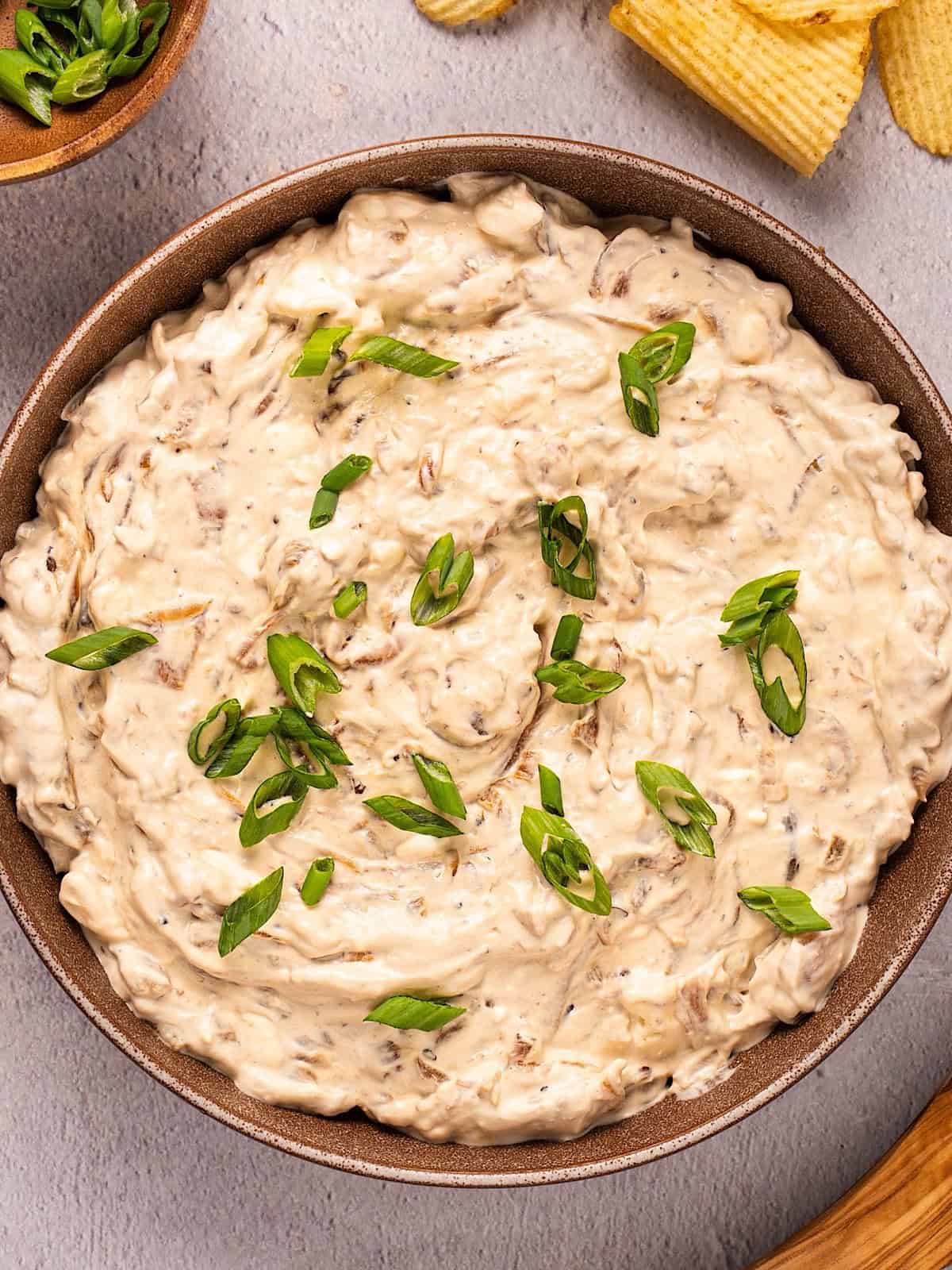 Caramelized onion dip with green onion garnish in a bowl plated next to chips and wooden spoon.