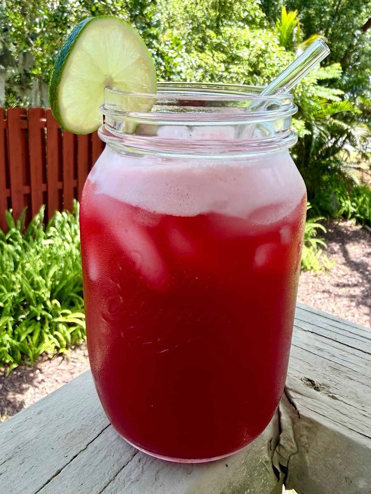 A mason jar sitting on a deck outside filled with cherry limeade, ice, and added garnish.