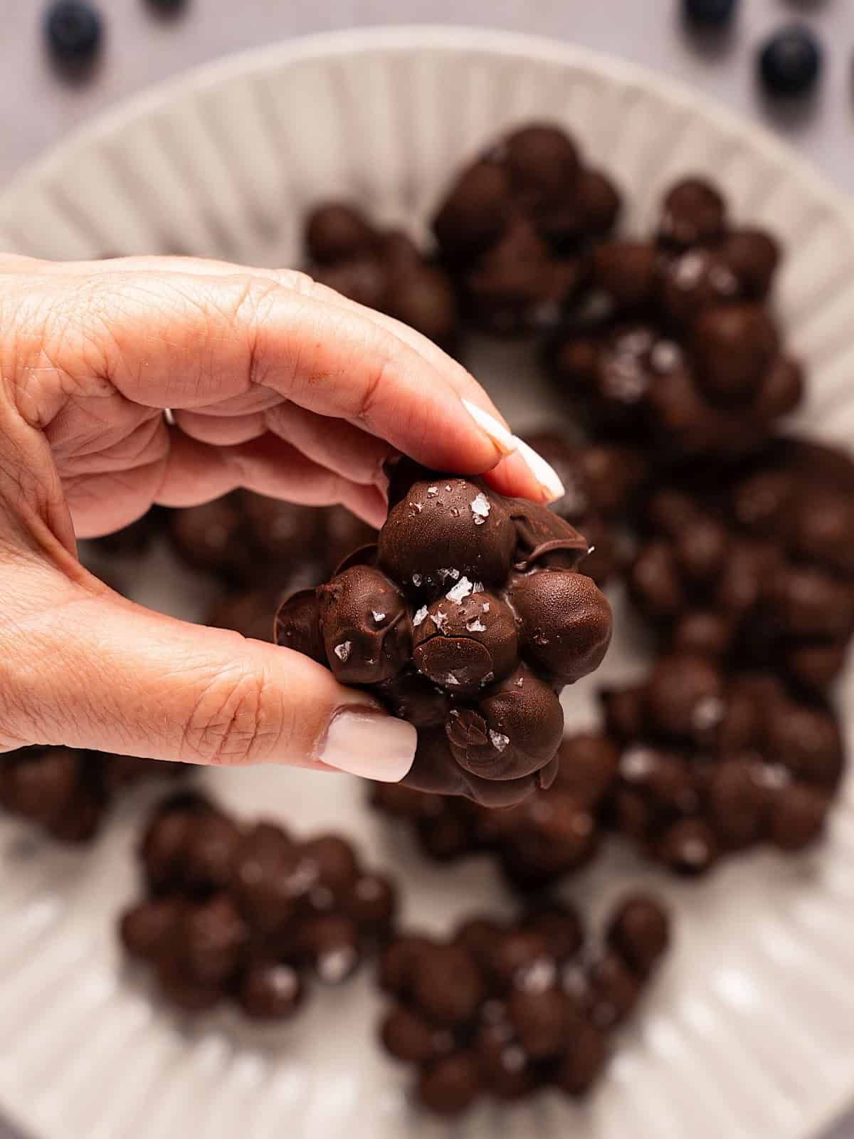 Holding a chocolate covered blueberry cluster with flaky salt above the full platter.