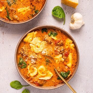 Two bowls of creamy Tuscan tortellini soup plated next to spinach leaves, parmesan rind, and garlic.