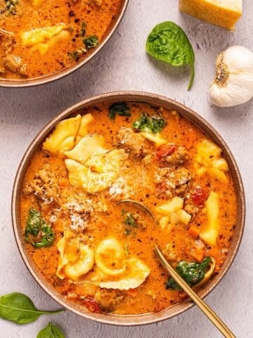 Two bowls of creamy Tuscan tortellini soup plated next to spinach leaves, parmesan rind, and garlic.