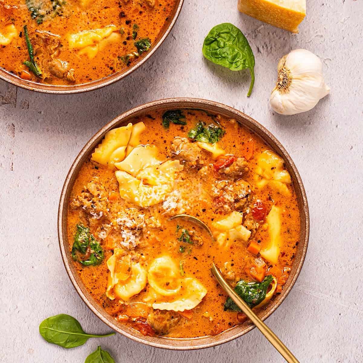 Two bowls of creamy Tuscan tortellini soup plated next to spinach leaves, parmesan rind, and garlic.