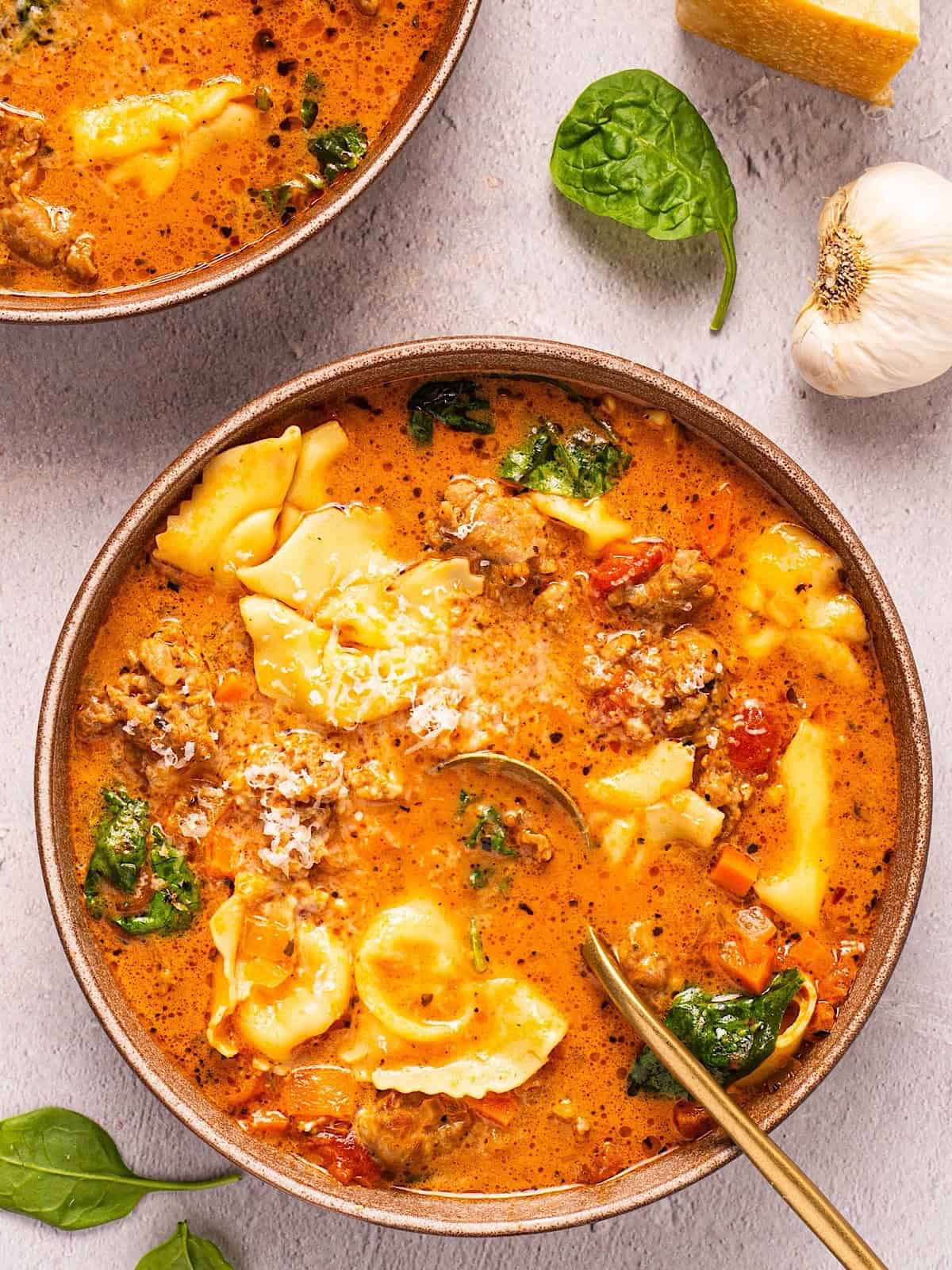 Two bowls of creamy Tuscan tortellini soup plated next to spinach leaves, parmesan rind, and garlic.