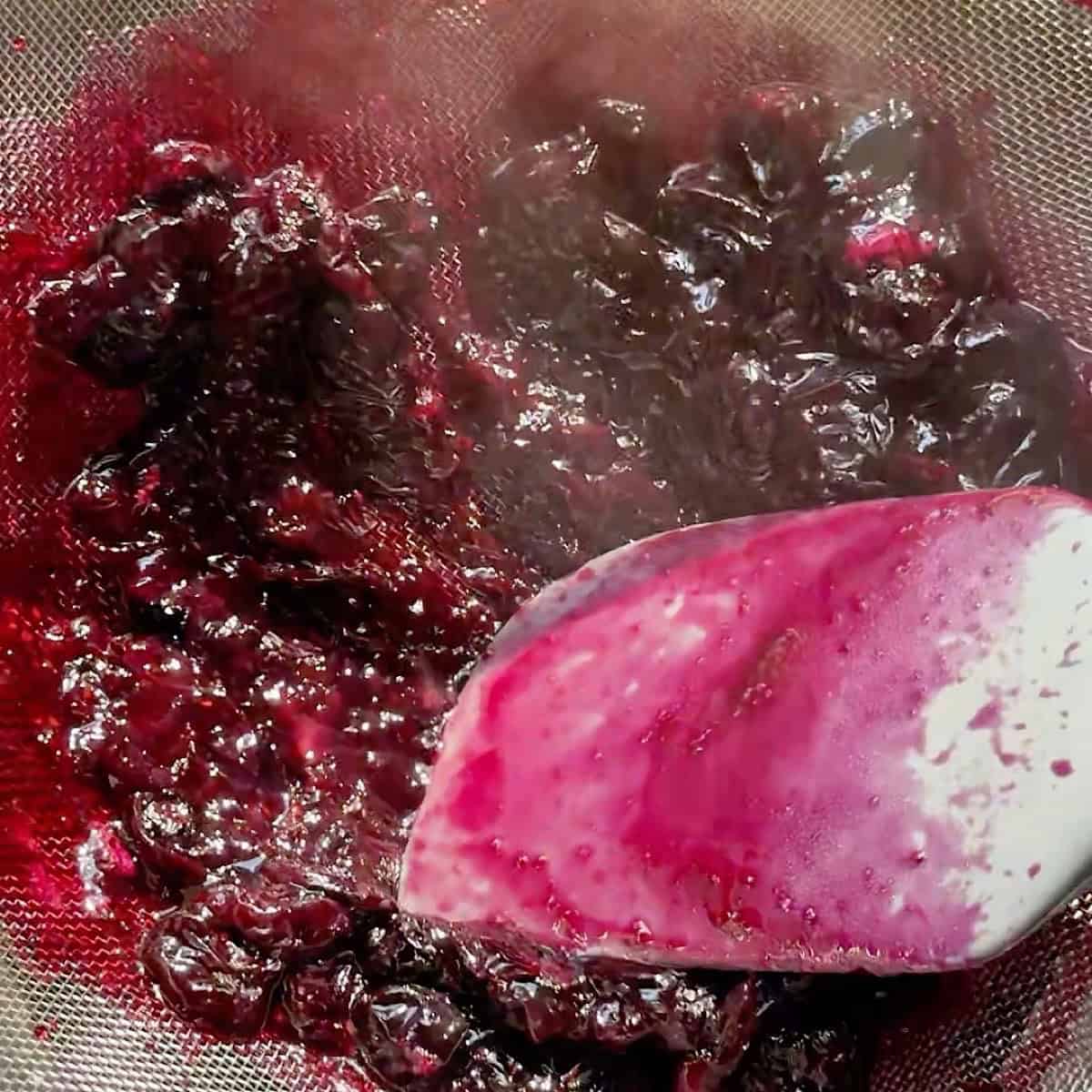 Straining the blueberries in a fine mesh sieve over a bowl and pressing down with a rubber spatula to extract juices.