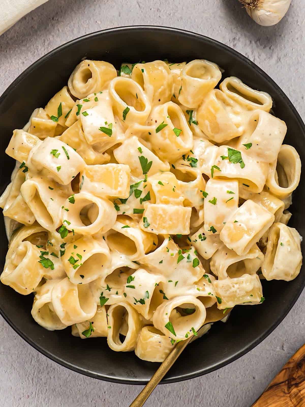 Alfredo coated pasta in a black bowl with parsley garnish next to linen and garlic bulb.